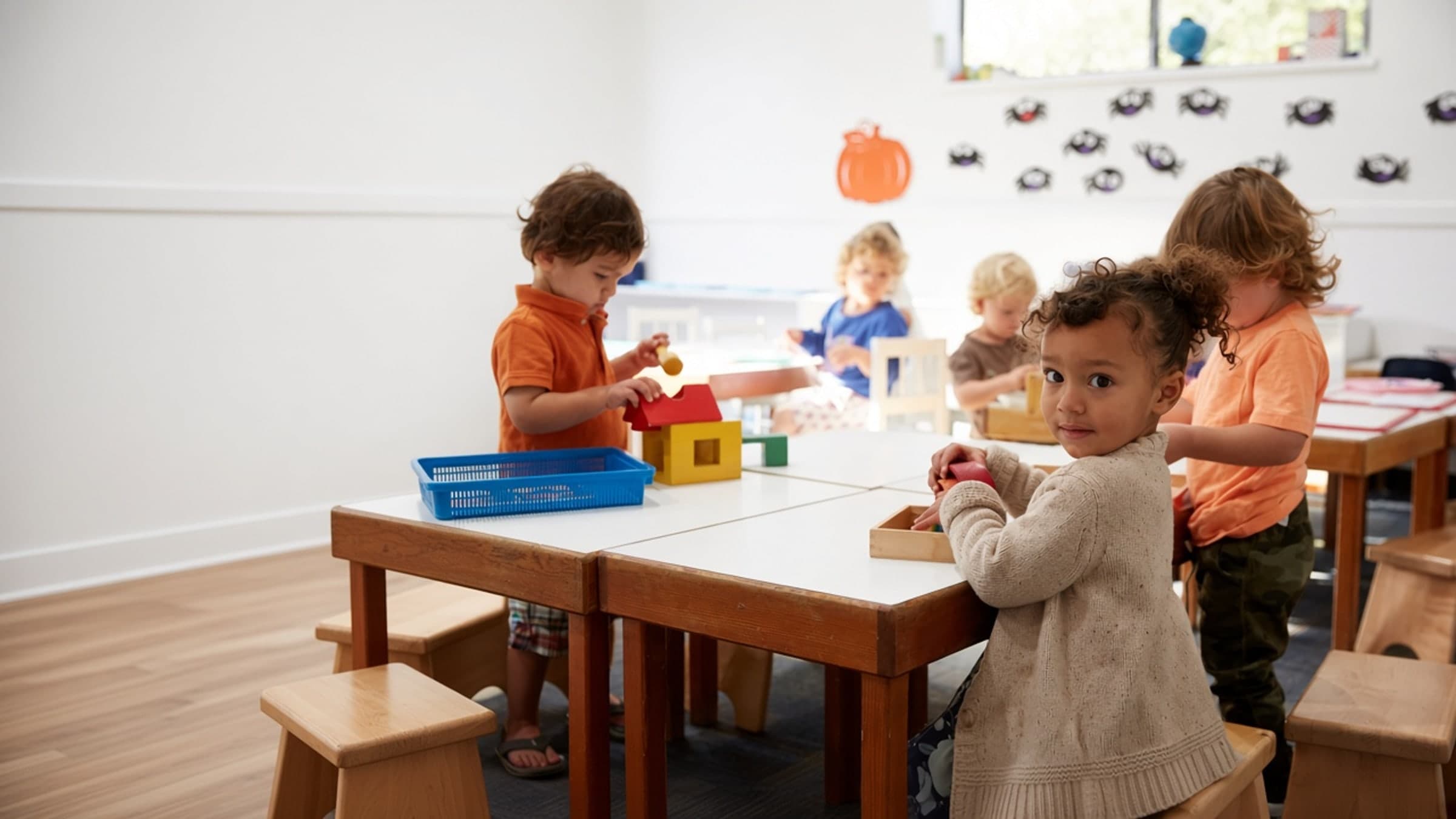 A preschooler looks toward the camera while classmates work at wooden tables